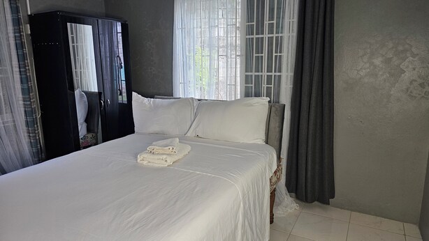 Una cama bien hecha con sábanas blancas y una toalla doblada, un armario de color oscuro y cortinas de encaje que dejan pasar la luz natural.