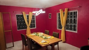 Sala de jantar com mesa de madeira posta para quatro pessoas, duas janelas com vidro gradeado e uma luminária pendente moderna.