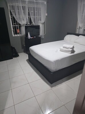 A hotel room with a bed, a nightstand, a TV, and a window with sheer curtains. - Relax & Unwind in Our Tranquil Space at Jamaica b&b rentals (Montego Bay)