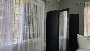 Un dormitorio con un ventanal cubierto por cortinas translúcidas, un armario negro y un almohadón blanco sobre la cama.