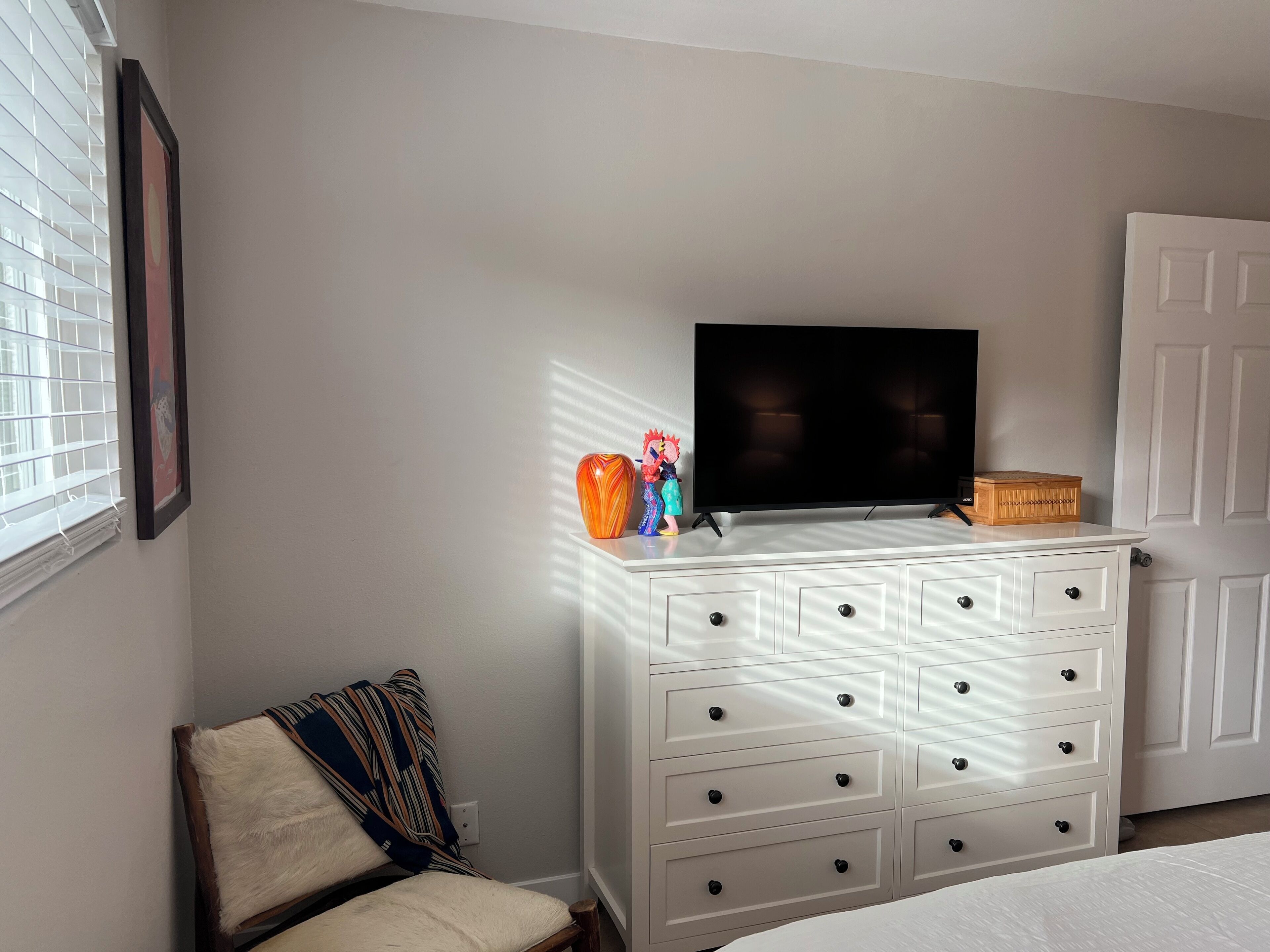 A bedroom with a bed, a dresser with drawers, a television on top, a window with blinds, and a door.