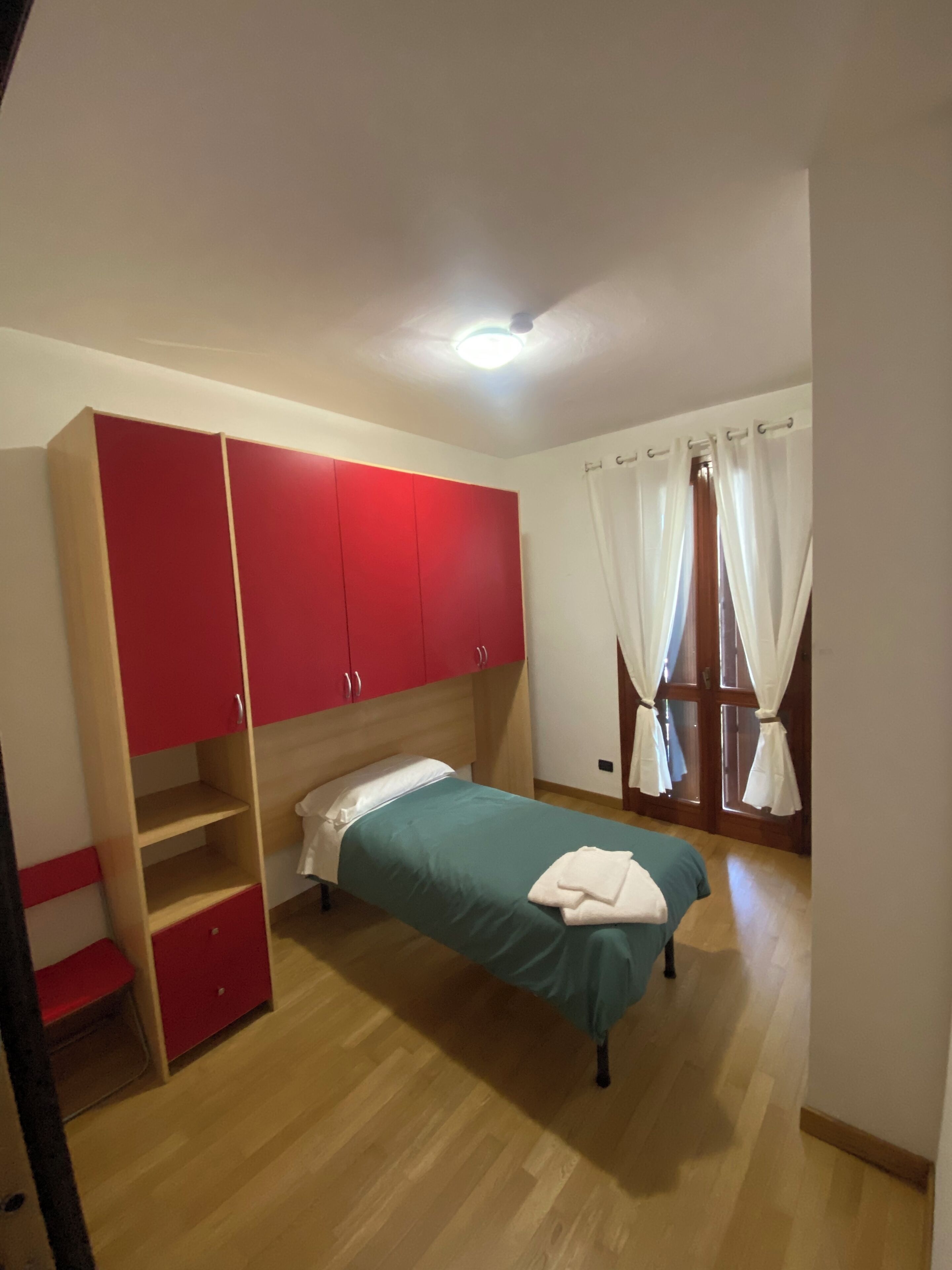 Ein Schlafzimmer mit einem Bett, einem roten Kleiderschrank, Holzboden und einem Fenster mit weißen Vorhängen.