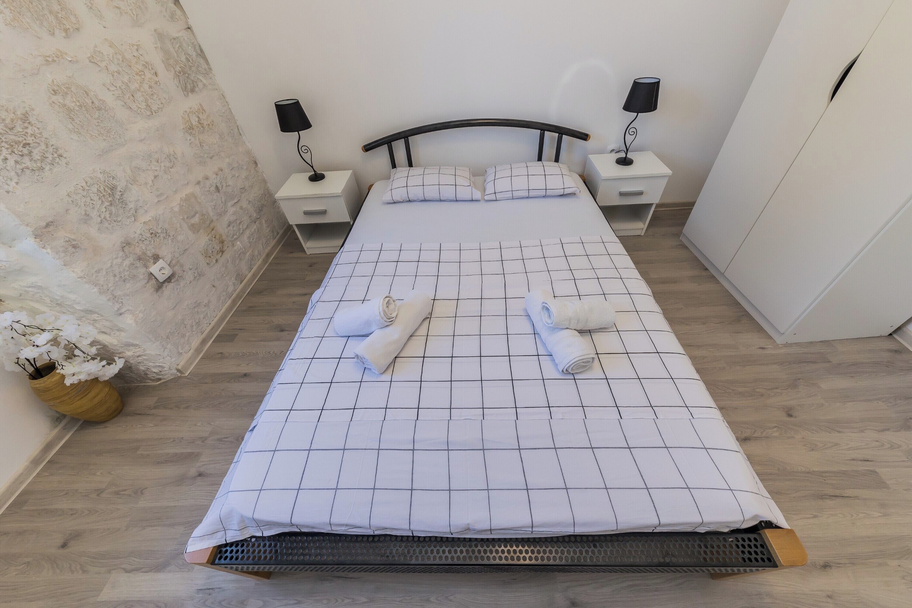 A neatly made bed with a grid-patterned bedspread, two bedside tables with lamps, a stone wall, and a wooden floor.