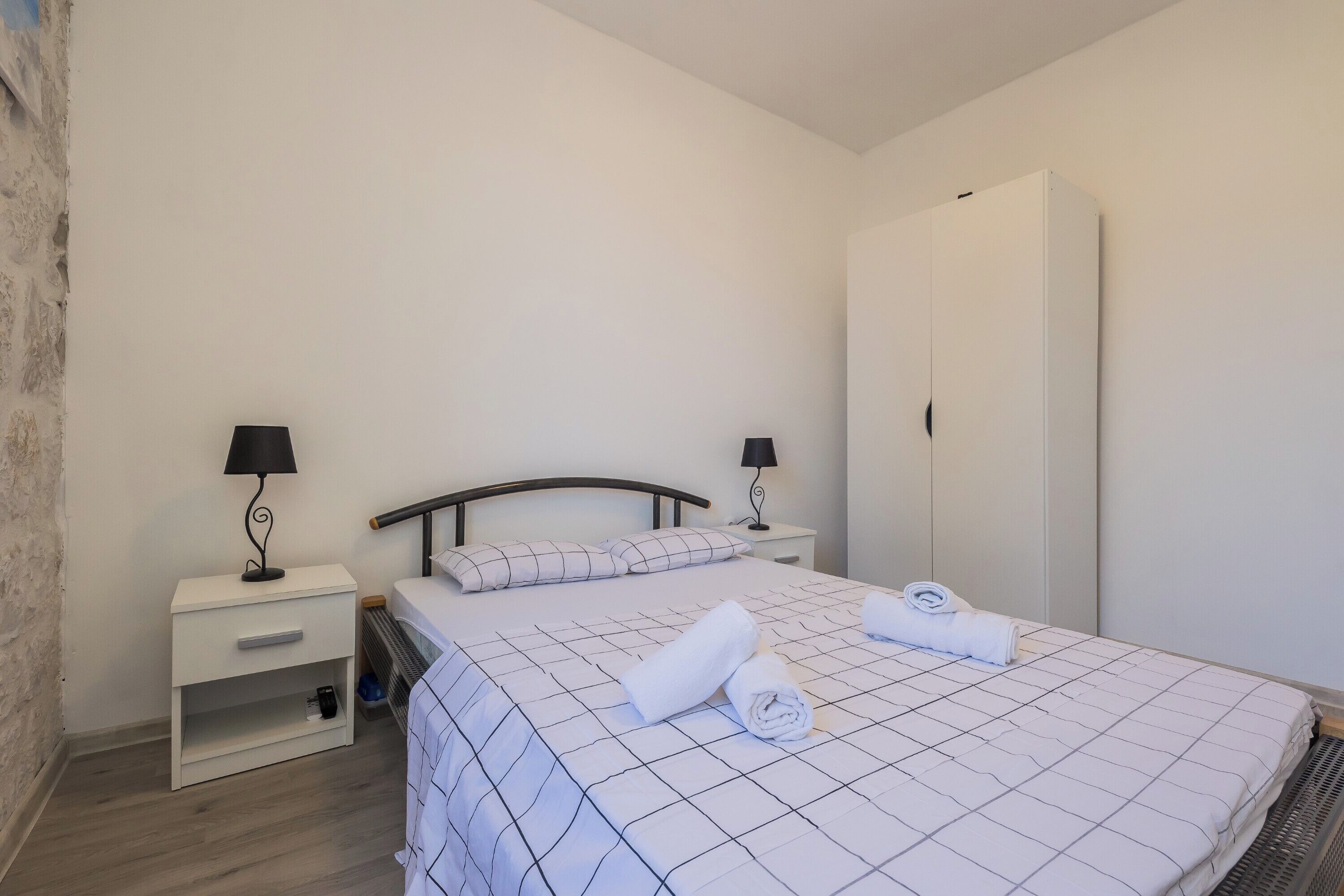 A neatly made bed with a checkered bedspread and rolled towels, a nightstand with a lamp, and a wardrobe in a simple, clean room.
