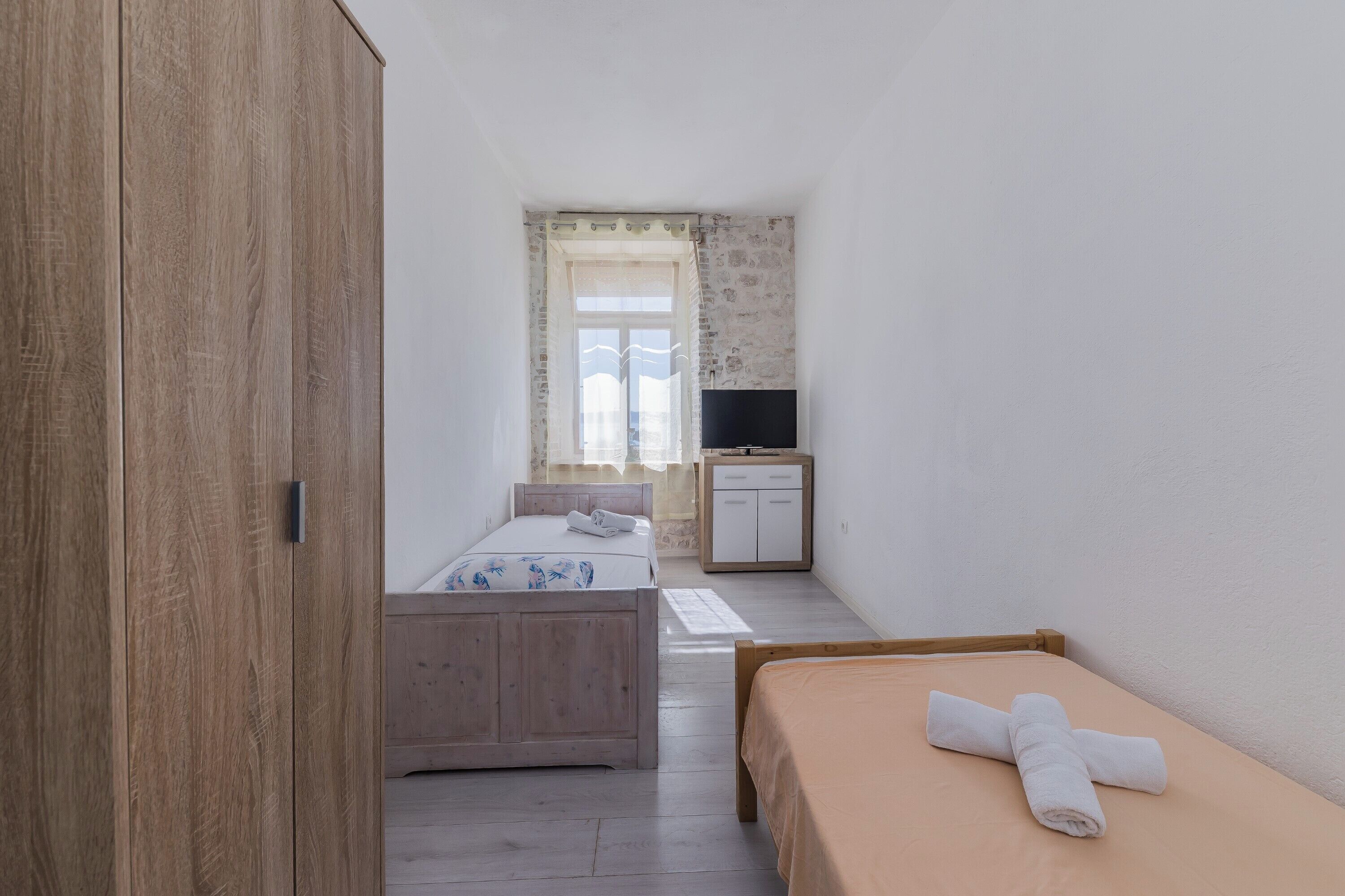 Un dormitorio con un armario de madera, una cama con edredón blanco, una mesita de noche, un pequeño mueble y un televisor.