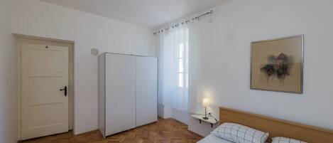 A hotel room with a wooden floor, a bed with white bedding, a nightstand with a lamp, a door, and a window with sheer curtains.