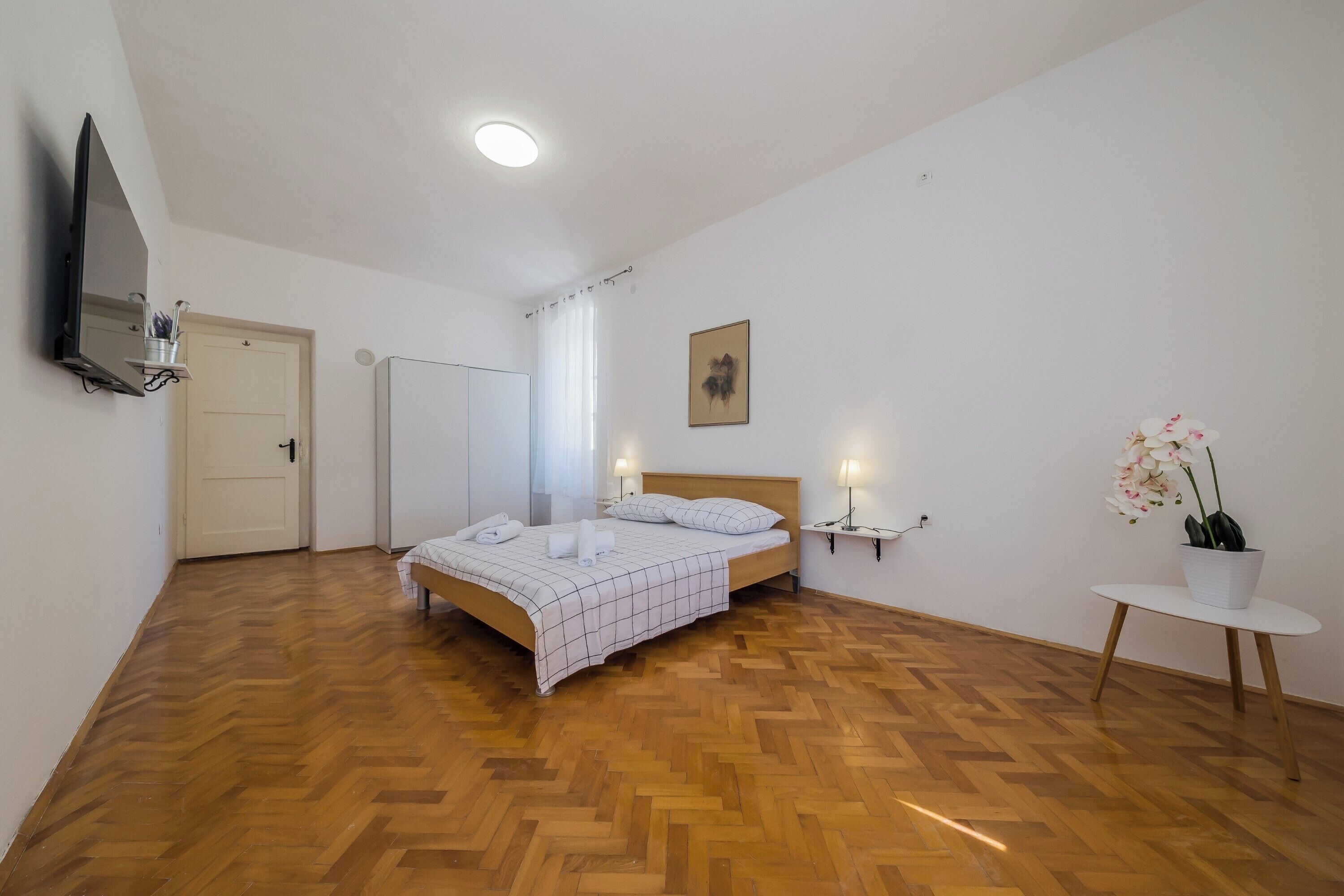 Quarto com piso de madeira em padrão herringbone, cama com colcha quadriculada branca e cinza, mesa de cabeceira com abajur e uma orquídea em vaso sobre uma pequena mesa.