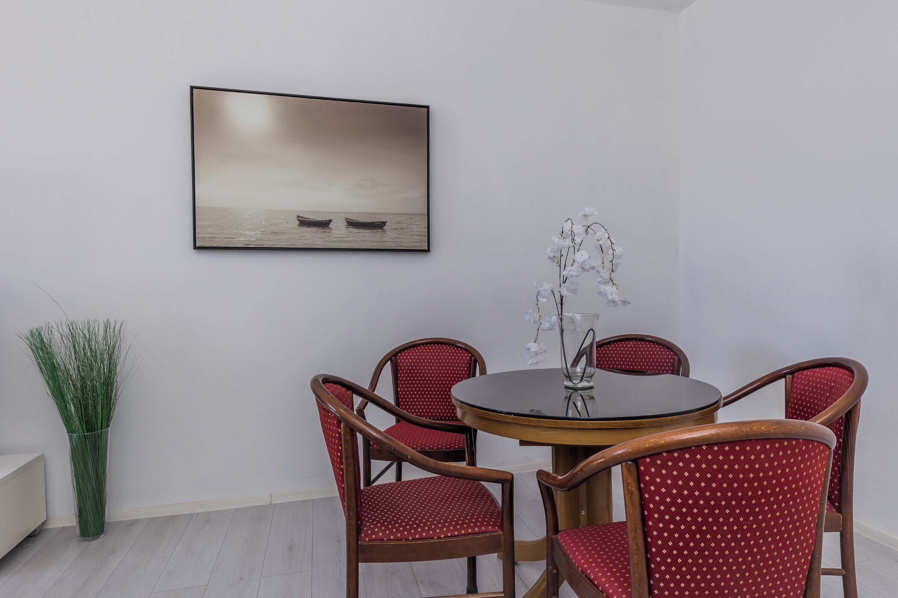 Un coin repas avec une table ronde, quatre chaises à l’assise rembourrée de couleur rouge, un vase avec des fleurs blanches et, au mur, un tableau représentant une mer calme.