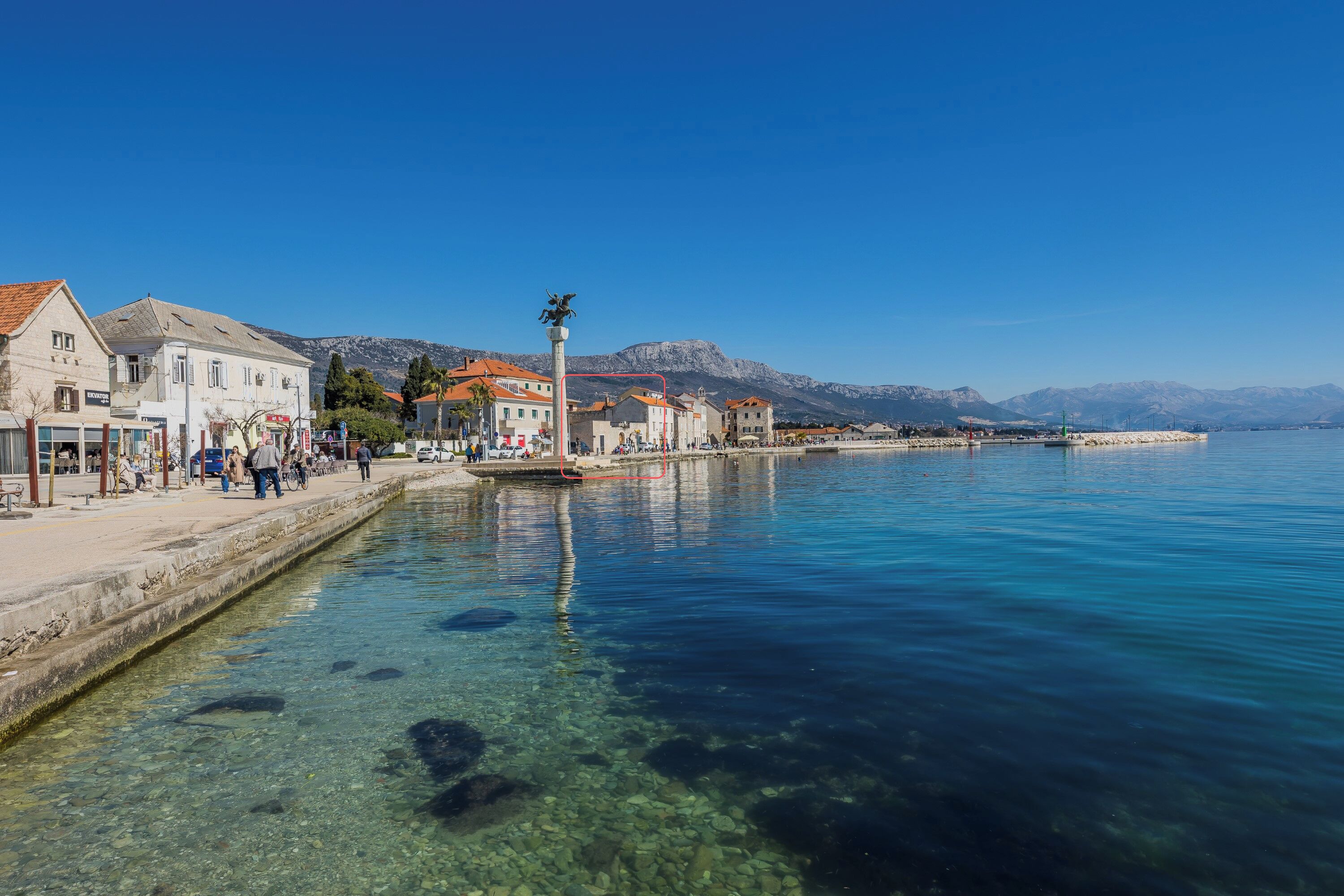 Un passeig marítim amb aigua cristal·lina, edificis i muntanyes al fons.