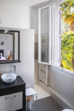 A modern bathroom with a white sink, a mirror, and a window with a view of palm trees. - Maison Tournesol Pau Jurançon Gan (Gan)