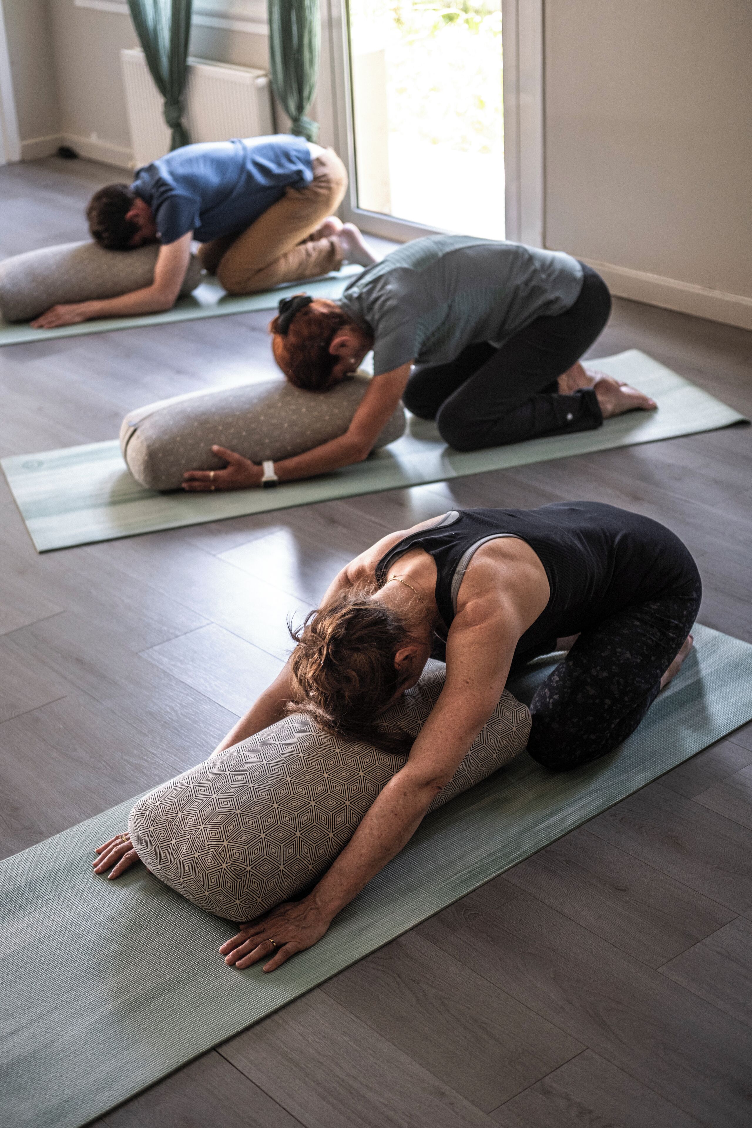 Tre personer dyrker yoga på måtter i et rum med trægulv og et vindue med gardiner.