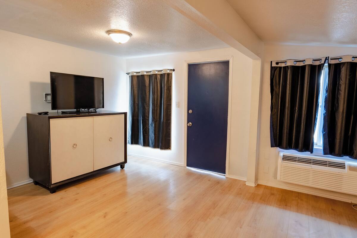 A room with a wooden floor, a TV on a cabinet, a door, and a window with curtains.