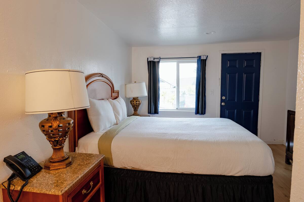 A hotel room with a bed, a nightstand with a lamp, a telephone, and a window with curtains.