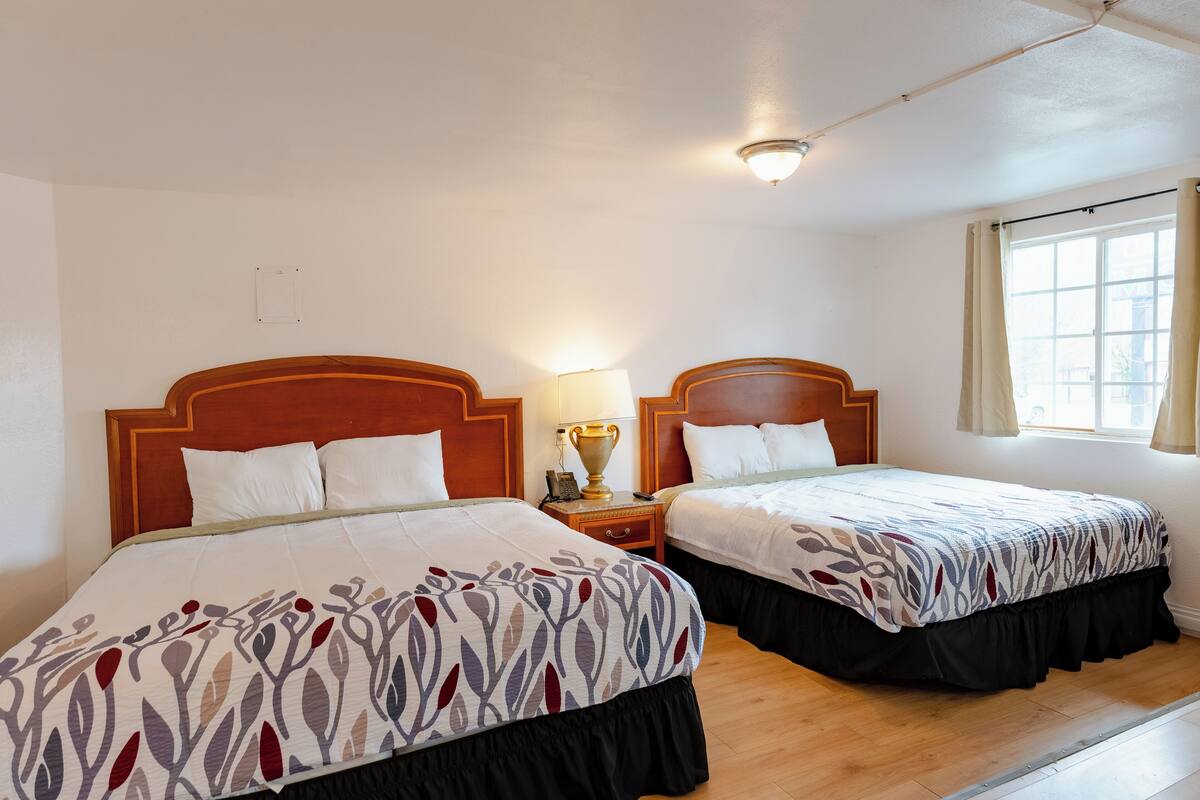 A hotel room with two beds, wooden headboards, a nightstand with a lamp, and a window with curtains.