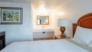 A hotel room with a bed, a bedside table with a lamp, a mirror above a sink, and a framed picture on the wall.