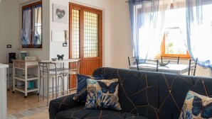A living room with a dark sofa, two butterfly-patterned cushions, a dining area with a table and chairs, and a kitchen area with a coffee machine and shelves.