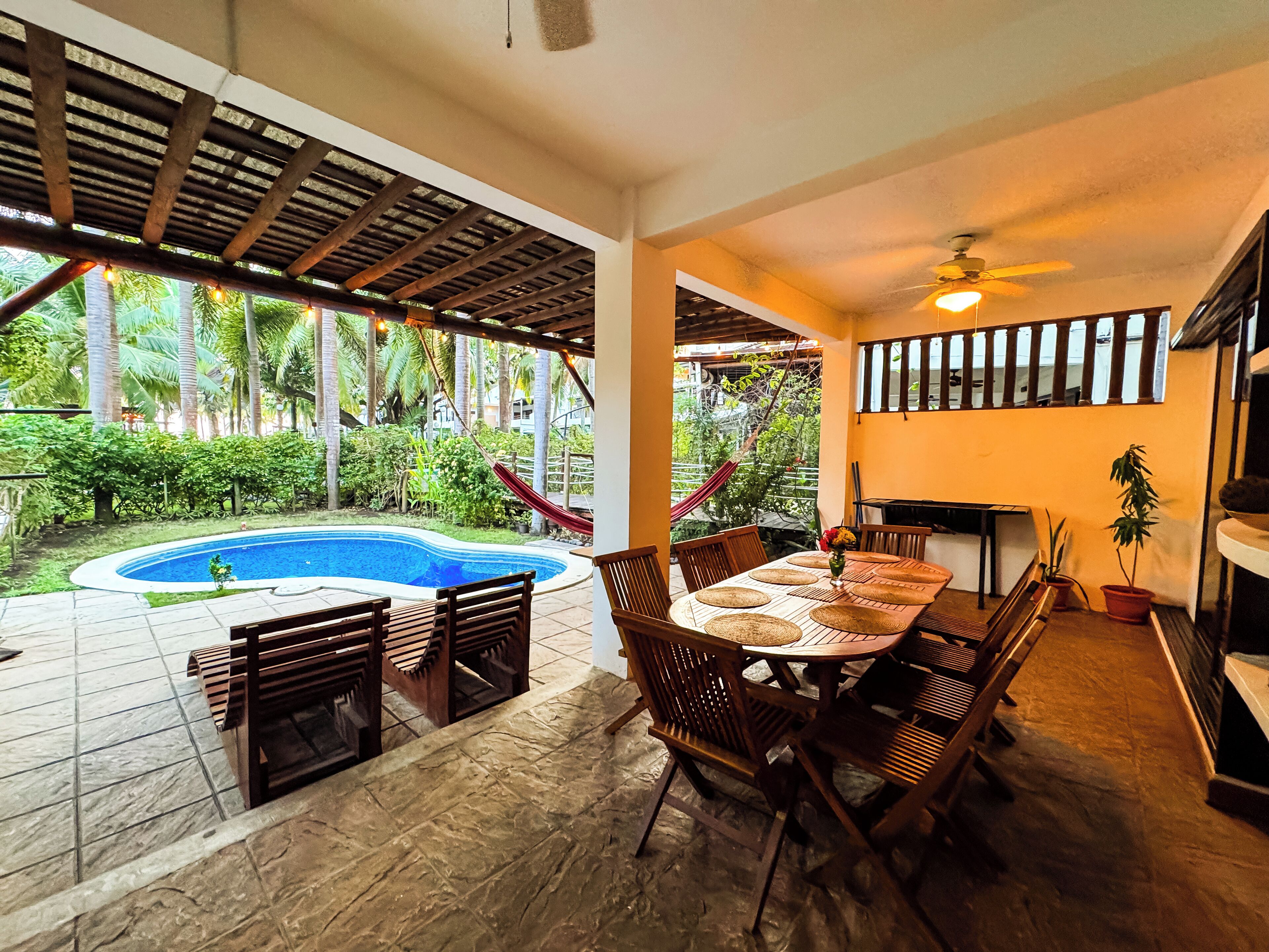 A covered outdoor dining area with a pool, wooden furniture, and a view of the surrounding greenery.