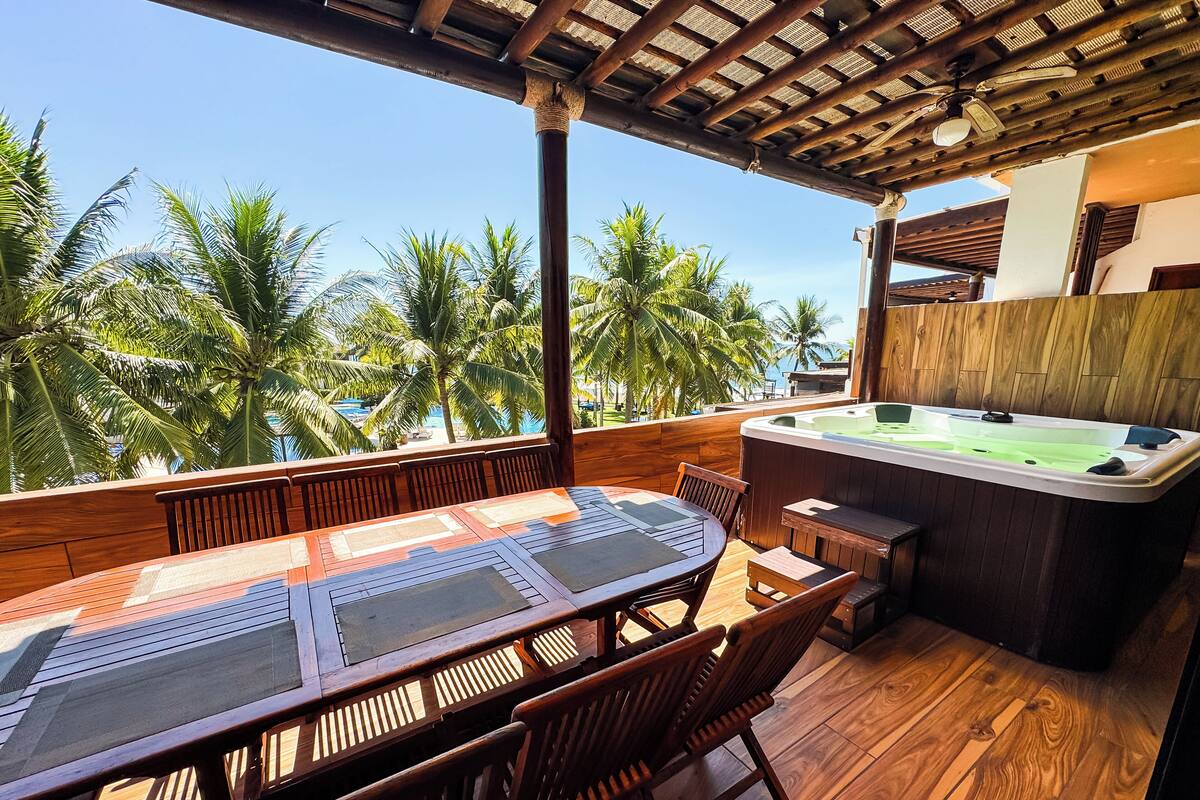 Een houten terras met een jacuzzi, een tafel en stoelen, met uitzicht op palmbomen en een heldere lucht.