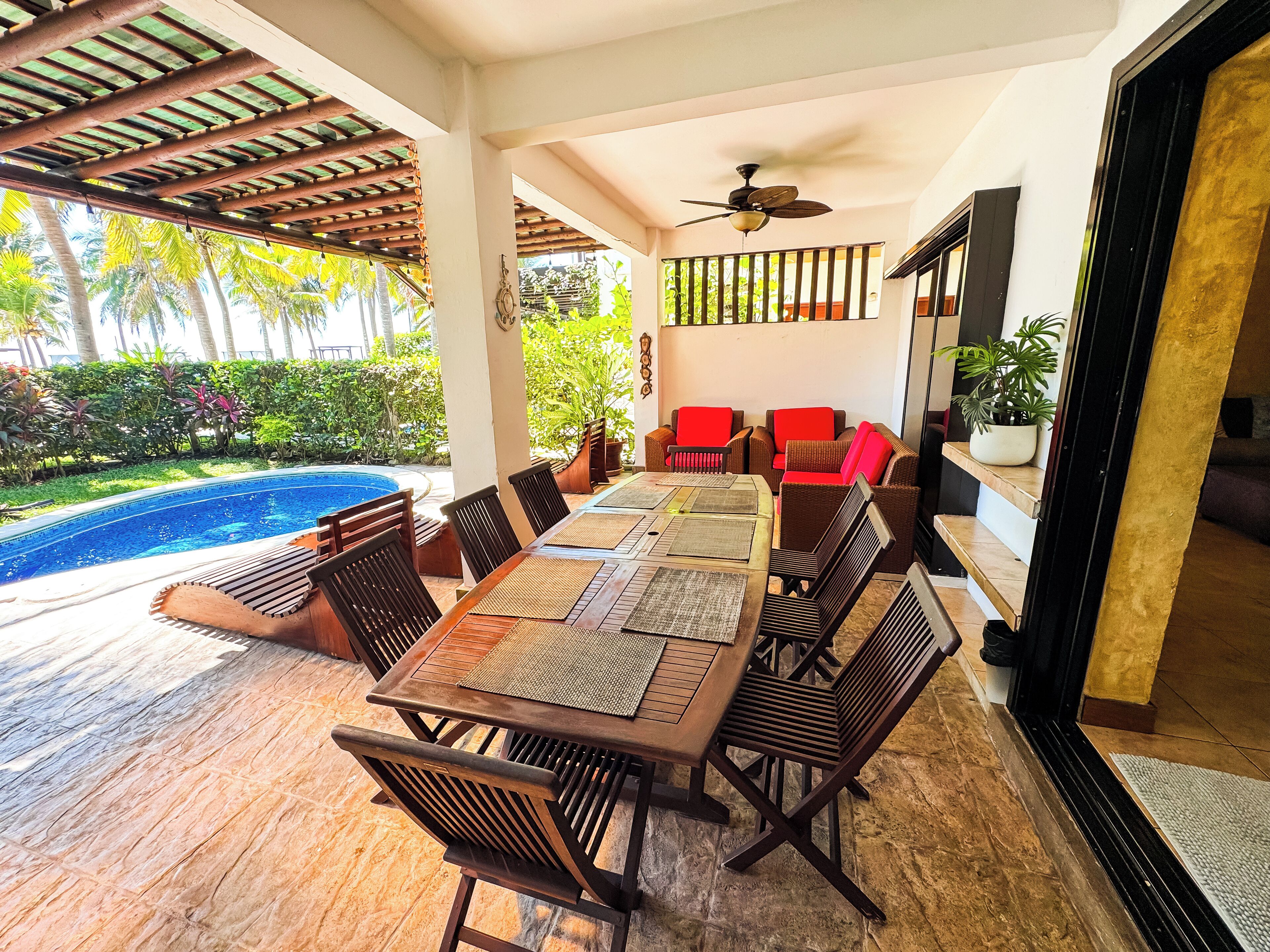 A covered outdoor dining area with a pool, wooden furniture, and a ceiling fan.