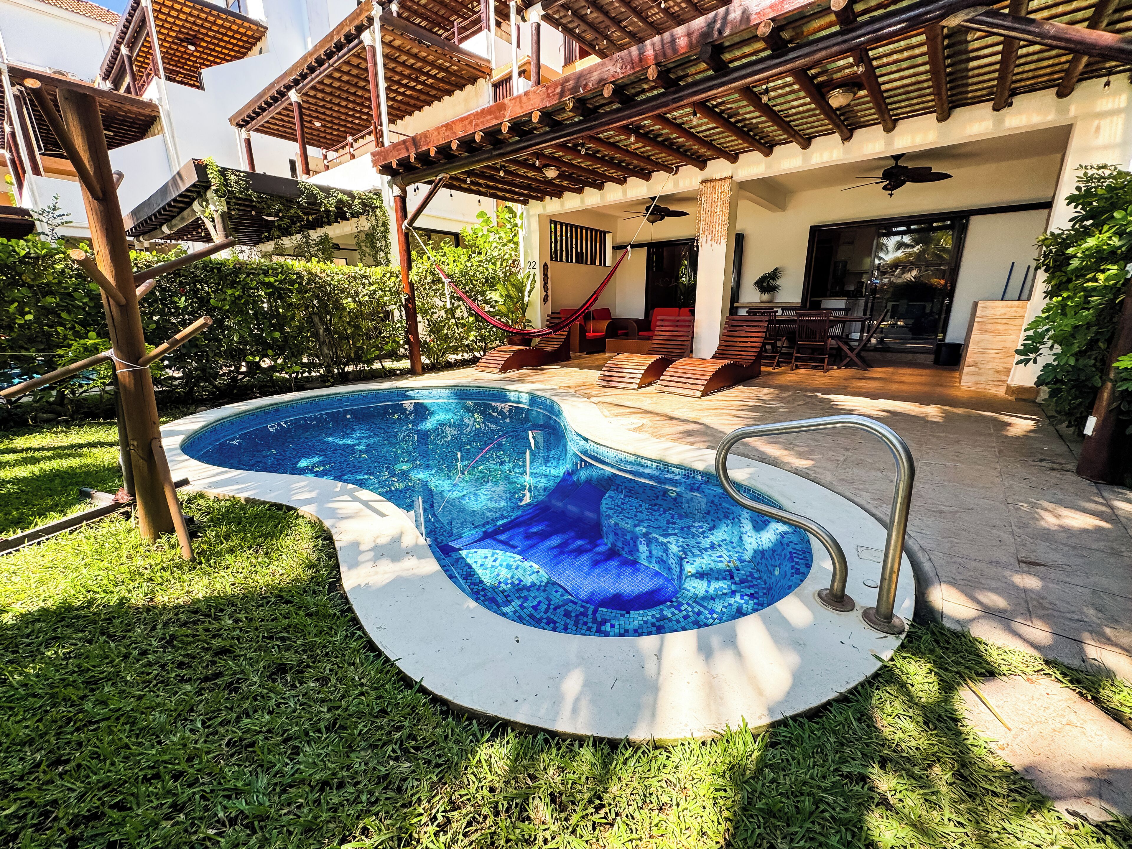 A swimming pool with a tiled deck, surrounded by grass and a wooden pergola.