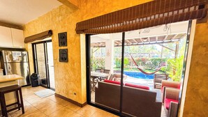 A kitchen with a refrigerator, a trash bin, and a sliding glass door leading to an outdoor seating area with a pool and hammock. - Villas Le Suisse Monterrico (Taxisco)