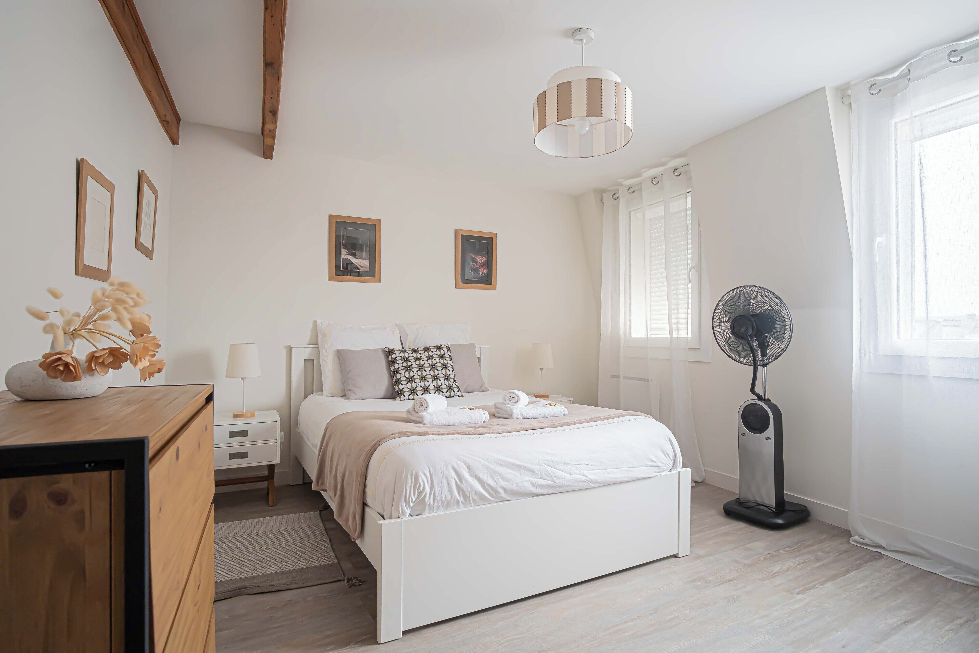 A bedroom with a bed, bedside tables, a standing fan, and a window with curtains.