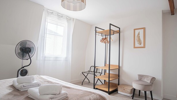 Habitación de hotel ordenada con cama, ventilador de pie, estante de madera, una silla y una obra de arte con hojas enmarcada en la pared.