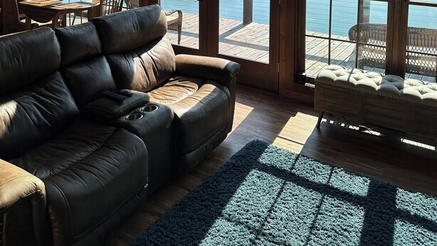A spacious living room with a leather sofa, a wooden dining table, and large windows overlooking a lake and dock. - Lakefront Cabin 3 bed, 3 1/2 bath with Private Dock (Sevierville)