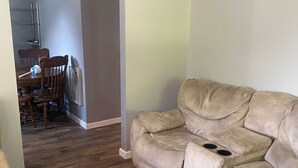 Un salon avec un canapé beige, un coin repas avec une table et des chaises en bois, et un couloir menant à une autre pièce.