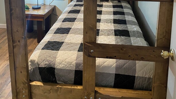 Un lit superposé avec une couverture à carreaux, un bureau en bois avec une lampe et une fenêtre avec des stores.