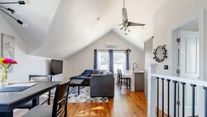 Ein modernes Wohnzimmer mit einer Couch, einem Esstisch und Stühlen. Es gibt einen Deckenventilator, einen Fernseher und ein Fenster mit Vorhängen.