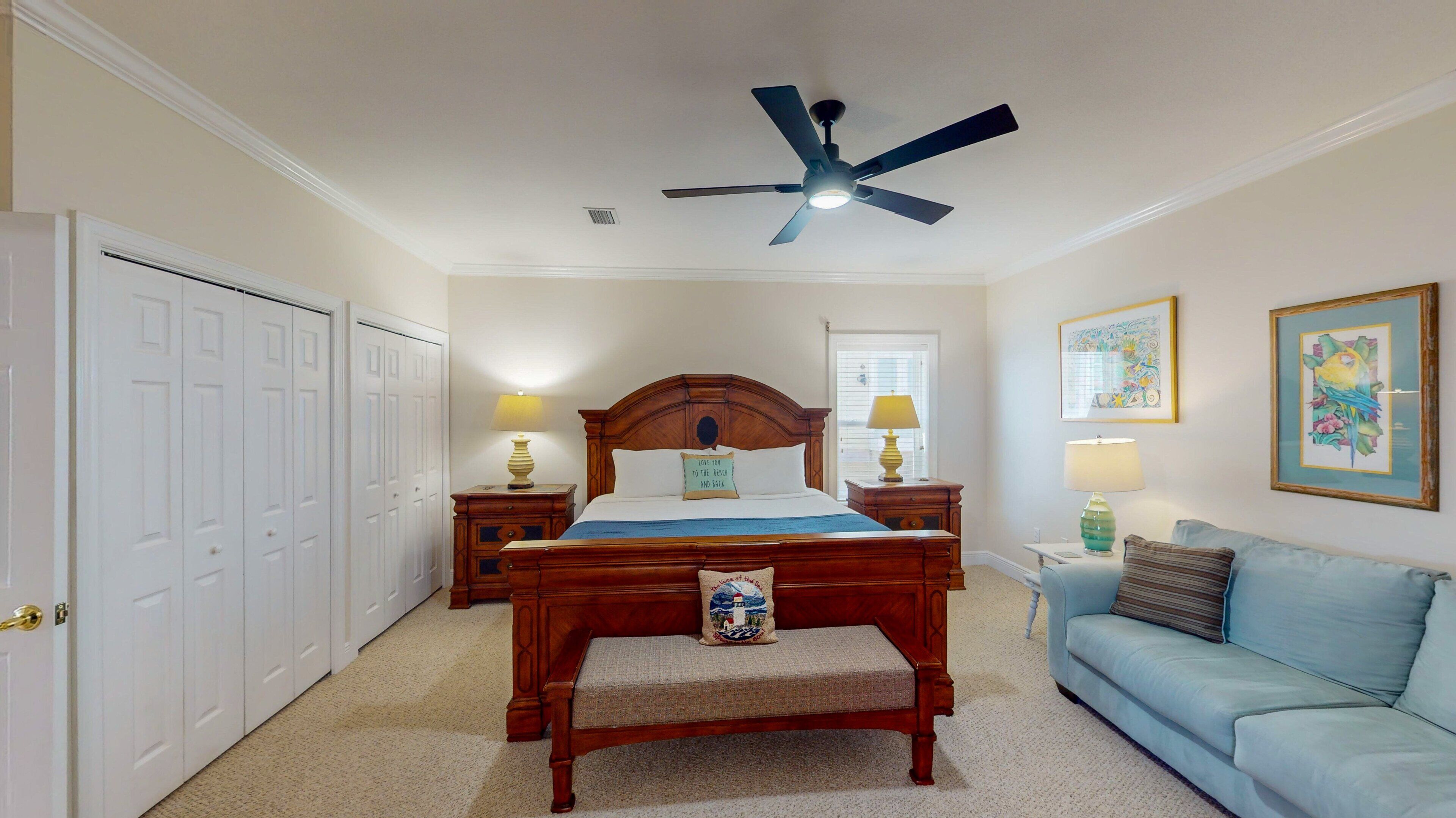 A bedroom with a bed, a ceiling fan, a nightstand, a bedside table, a couch, and a door.