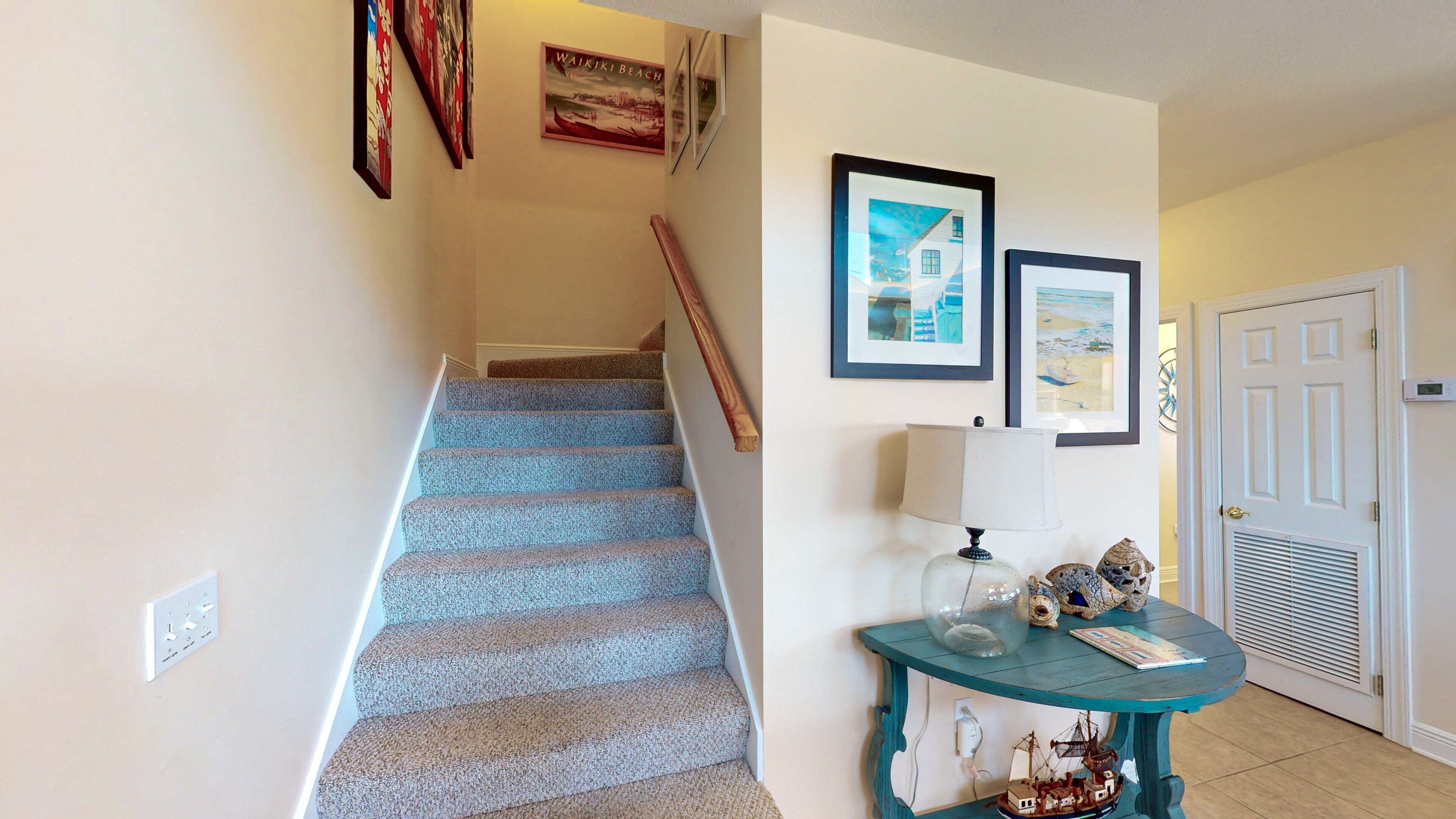 Un escalier recouvert d’une moquette bleue, une rampe en bois et un mur orné de tableaux encadrés et d’un thermostat.