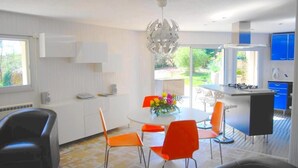 Een moderne keuken met een eethoek, met een ronde tafel met oranje stoelen, een wit kastje en uitzicht op de tuin door het raam.