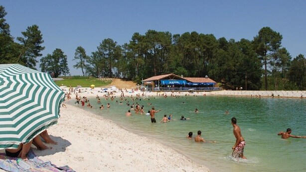 Een strand met mensen die zwemmen en zonnebaden, een gestreepte parasol en een gebouw met een blauw dak op de achtergrond.