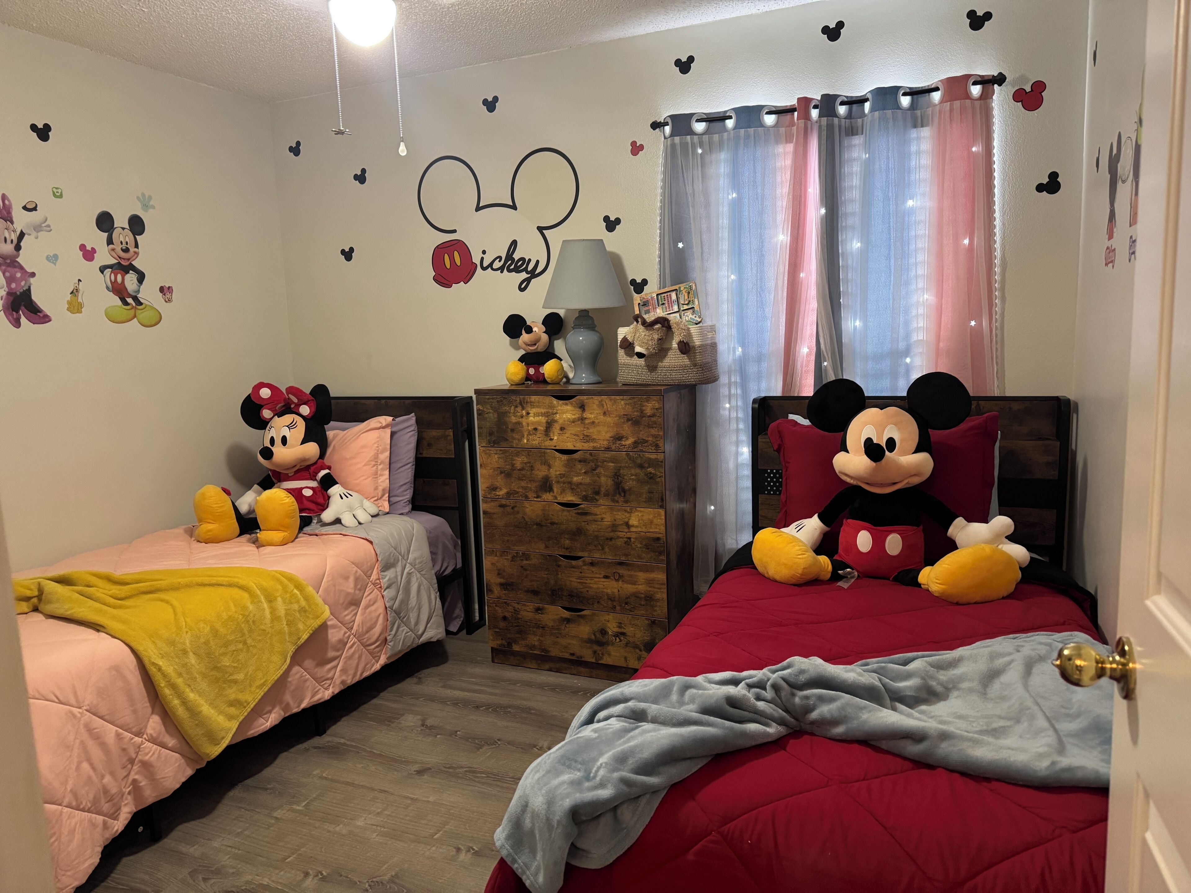A bedroom with two beds, each with a Mickey Mouse plush toy, a dresser, a lamp, and wall decals.