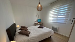 Un dormitorio moderno con una cama grande, una mesita de noche con lámpara, un escritorio con computadora y una ventana con persianas.