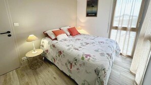 Un dormitorio con cama, mesita de noche, lámpara, un cartel en la pared y ventana con cortinas.