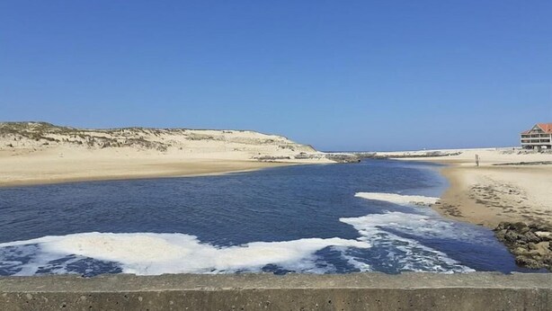 Paisaje costero con una playa, un edificio de techo rojo y un cielo azul despejado.