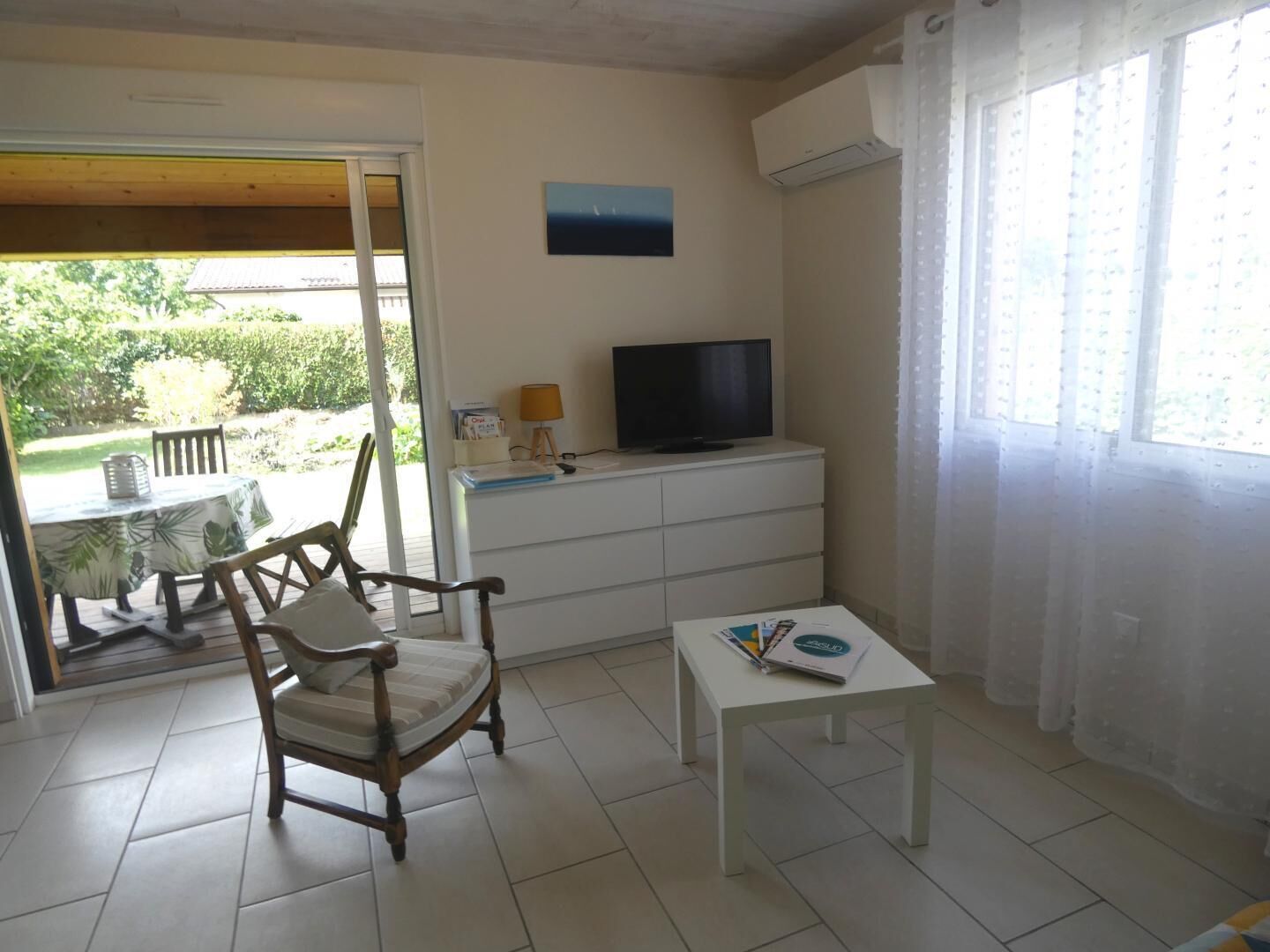 A room with a wooden chair, a small white table, a TV on a white cabinet, and a view of greenery outside.
