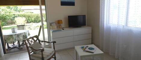 A room with a wooden chair, a small white table, a TV on a white cabinet, and a view of greenery outside.