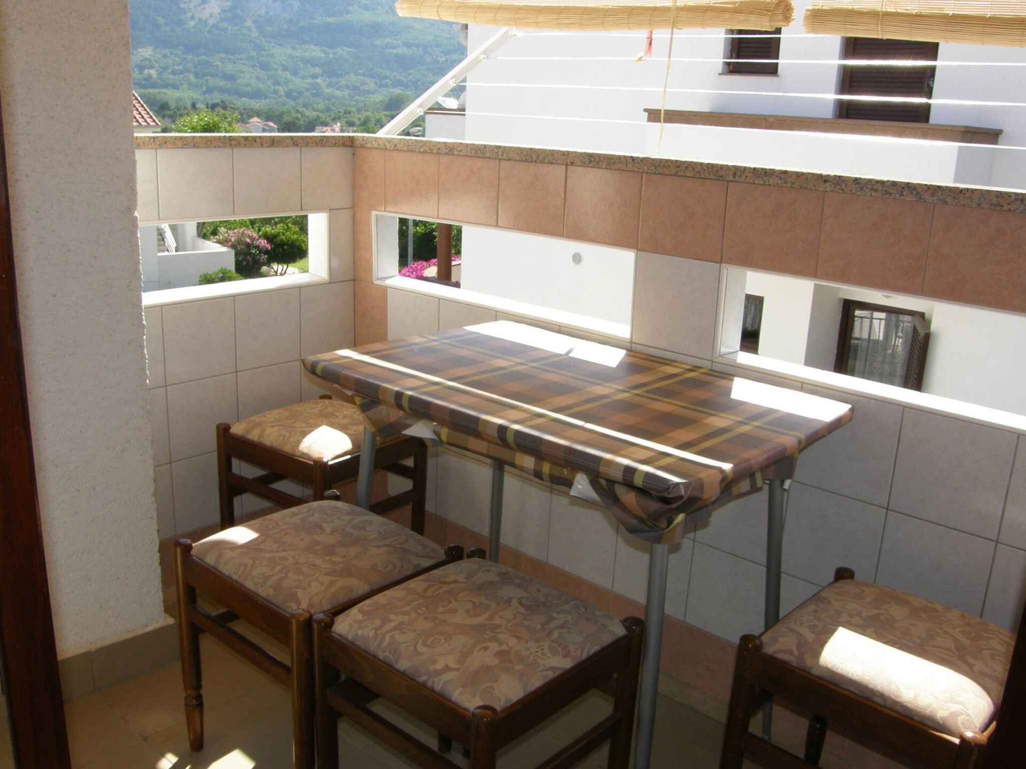 Pequena varanda com uma mesa e três cadeiras, vista para área verde e um edifício ao fundo.