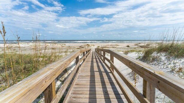 Een houten looppad dat naar een strand met duinen en schaarse begroeiing leidt.