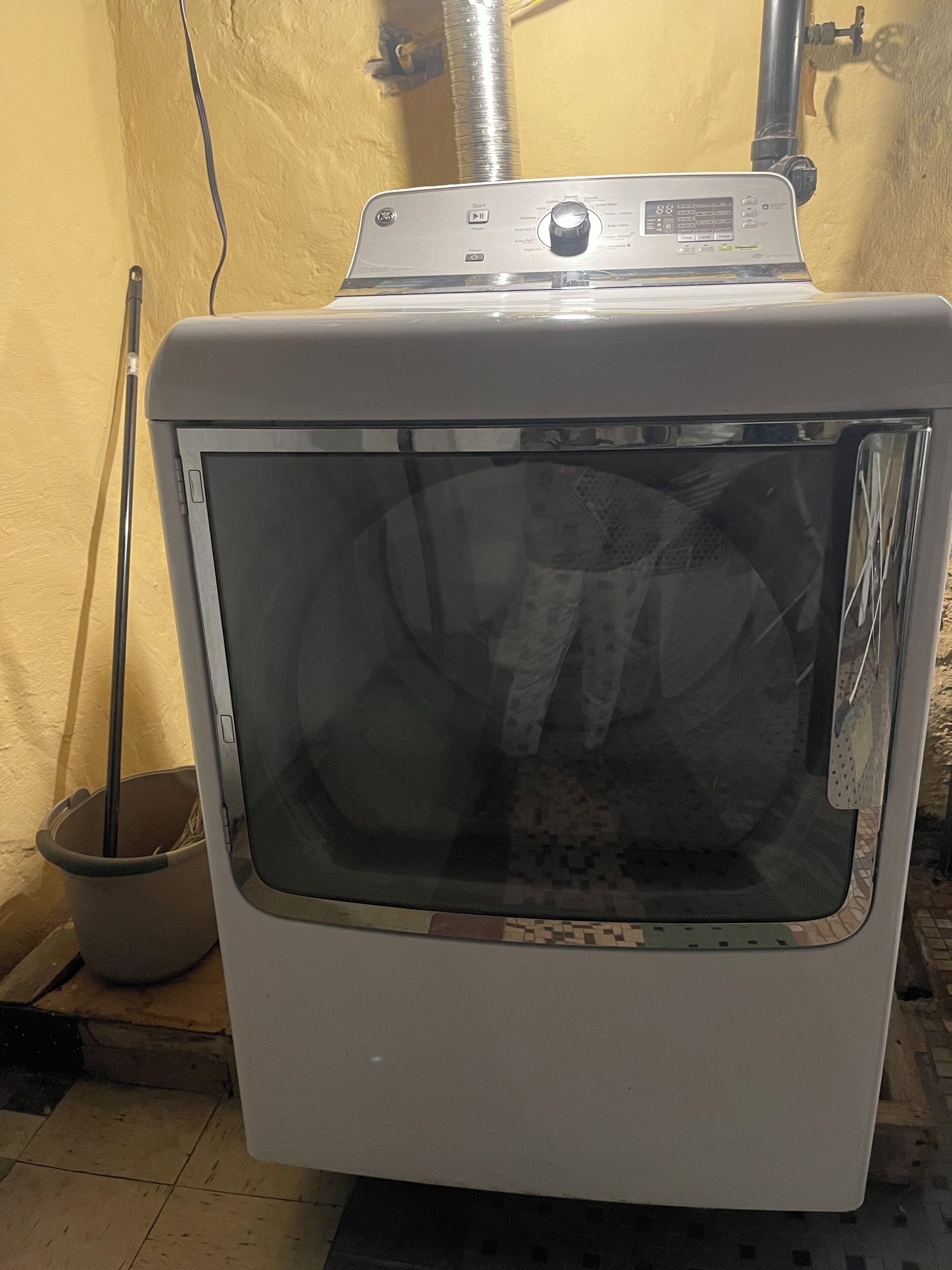 En hvid tørretumbler med kontrolpanel, en moppe og en balje på et flisegulv.