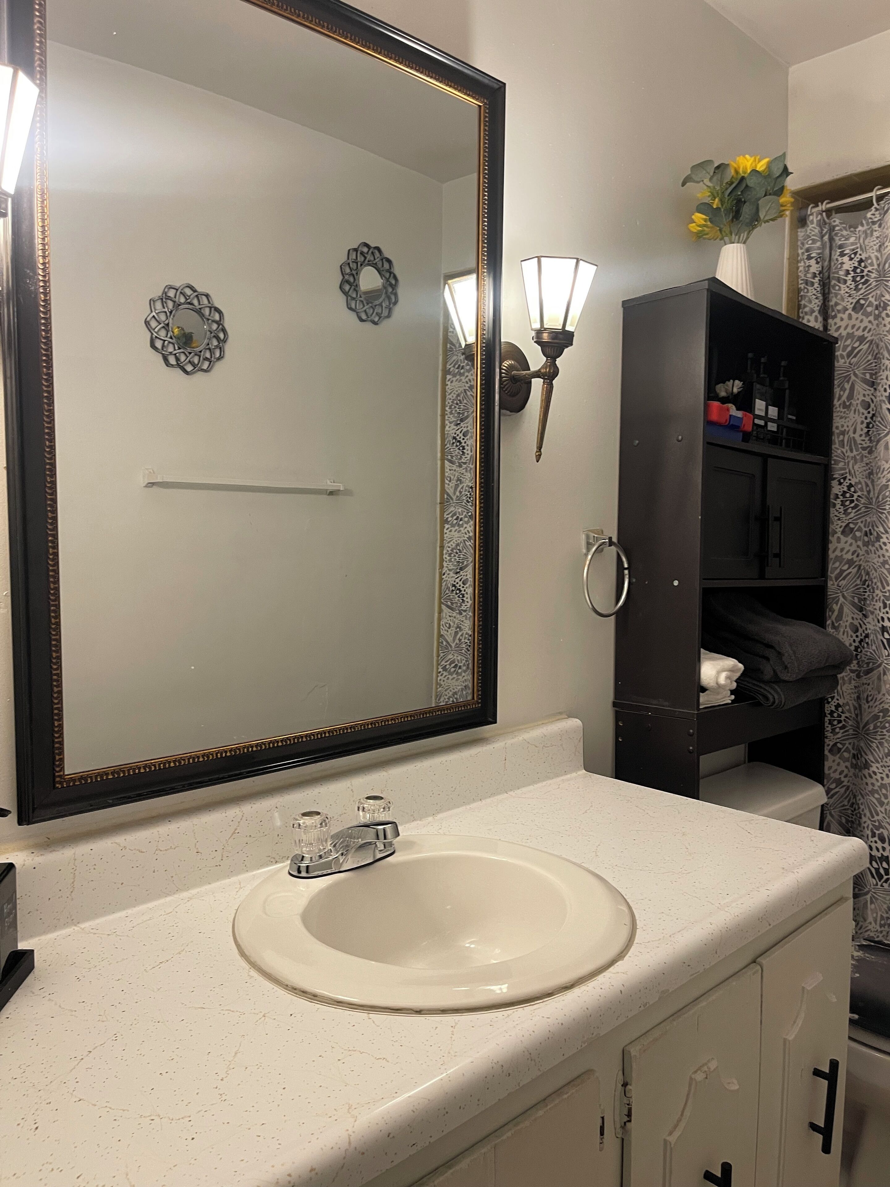 Un baño con un espejo grande, un lavamanos blanco, un mueble oscuro y una lámpara fijada a la pared.