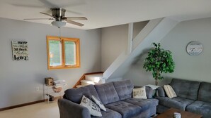 Un salon avec un canapé gris, une table basse en bois et un escalier. On y trouve une plante et une horloge murale.