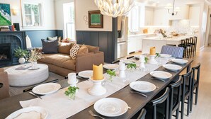 Ein moderner Essbereich mit einem langen Tisch, der für eine Mahlzeit gedeckt ist, ein gemütliches Wohnzimmer mit Kamin und eine Küche im Hintergrund.