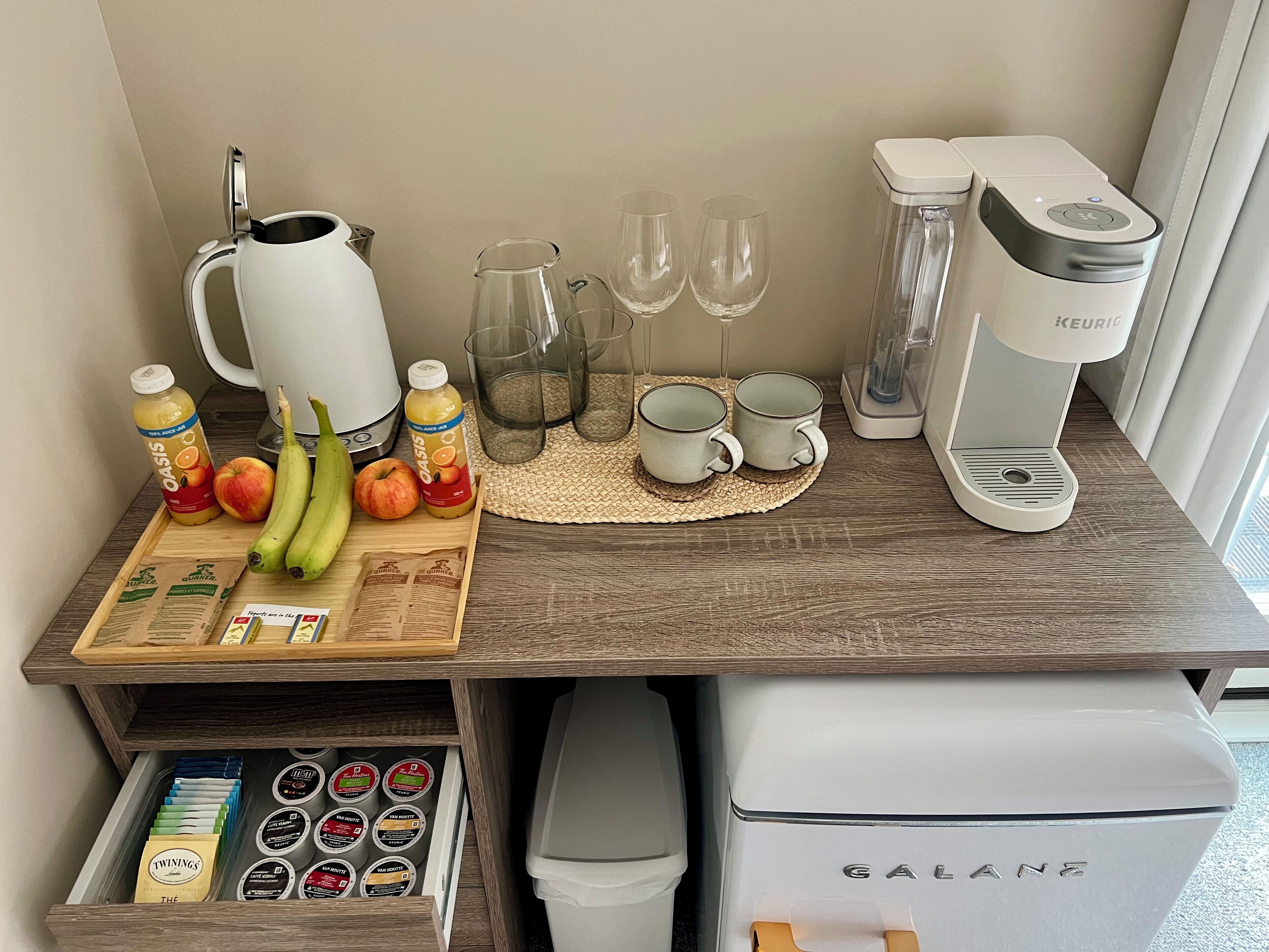 A kitchenette with a coffee maker, a kettle, a fruit tray, and a drawer with tea and coffee pods.