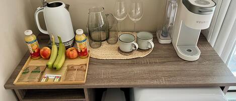 A kitchenette with a coffee maker, a kettle, a fruit tray, and a drawer with tea and coffee pods.