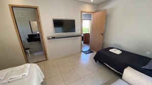 A bedroom with a bed, a TV mounted on the wall, a small desk, and a bathroom visible through an open door. - Villa Querência (Avare)